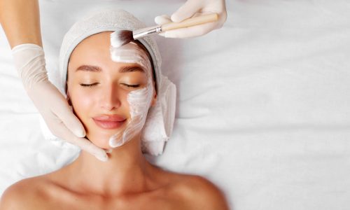 Beautiful young woman getting nourishing facial mask at spa salon, cosmetologist applying white mask with brush, top view, panorama with copy space. Facial beauty treatment concept